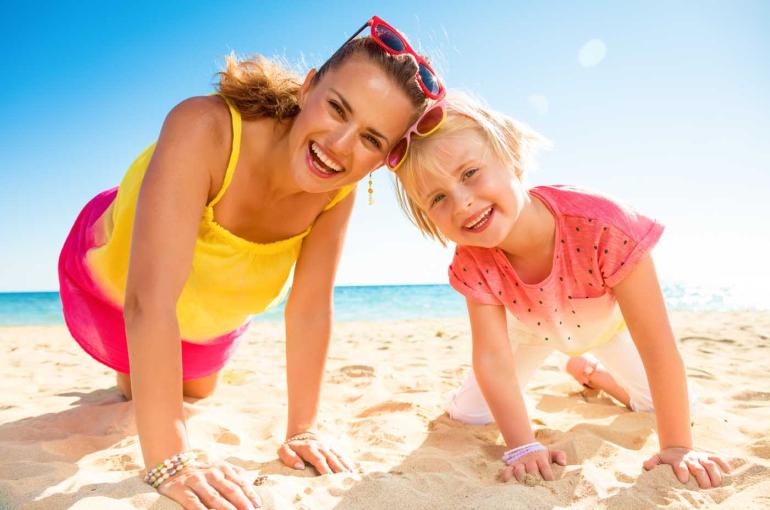 Madre e figlia sorridenti giocano felici sulla spiaggia sotto un cielo limpido.